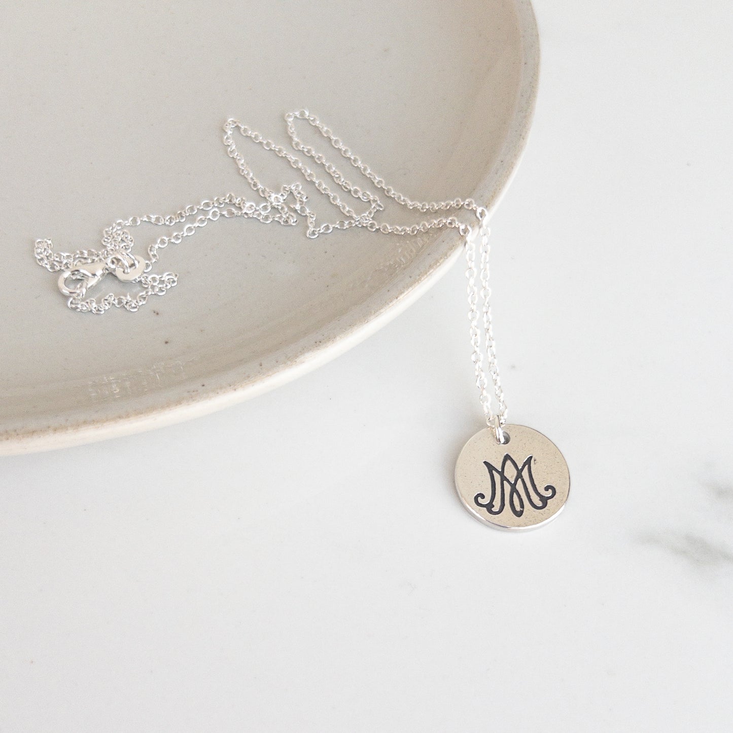 Silver necklace with a round pendant featuring letters "A" and "M" intertwined, for "Auspice Maria," on a white surface.