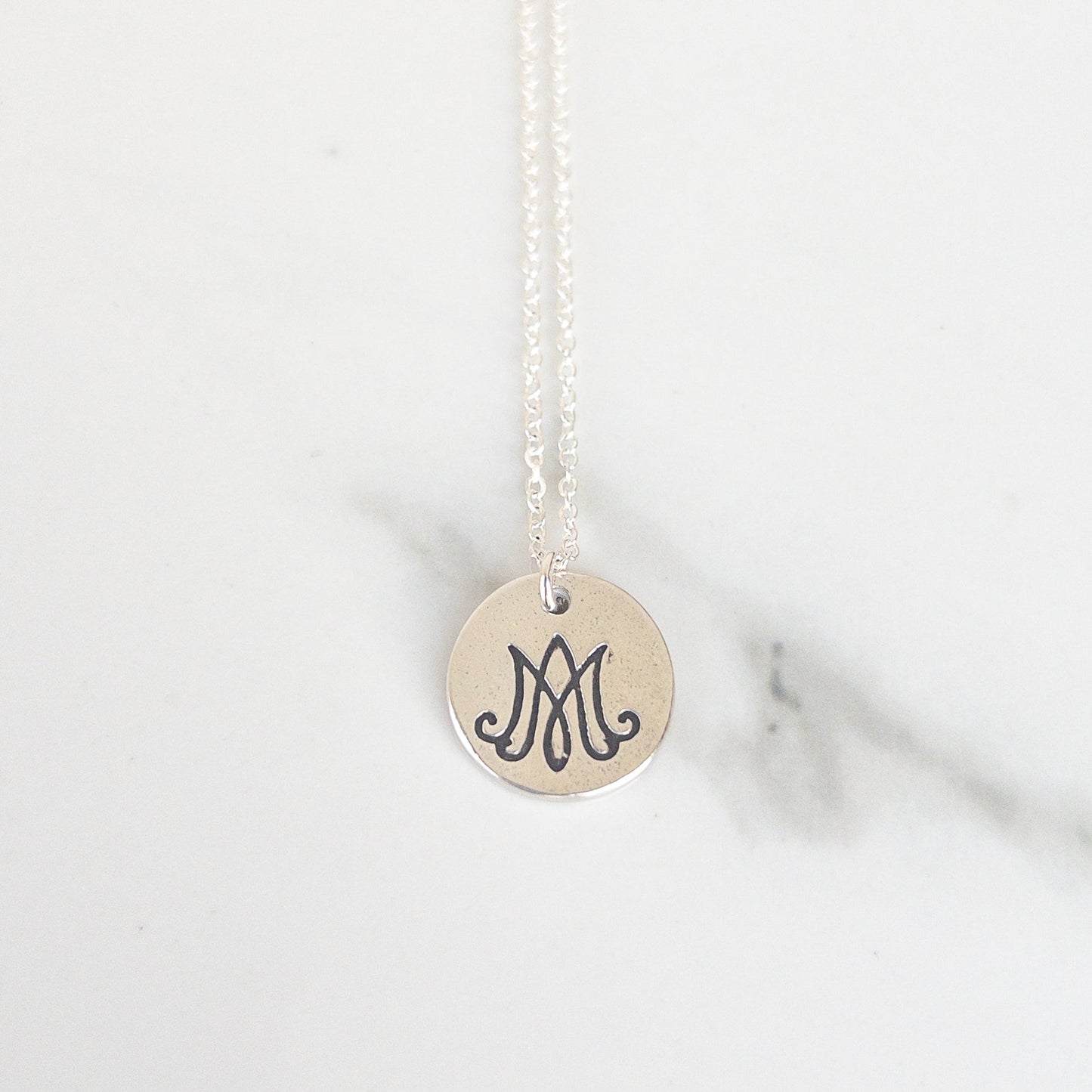 Close-up of round Marian monogram pendant in sterling silver, Auspice Maria symbol on white background. Necklace by Sagely Sparrow