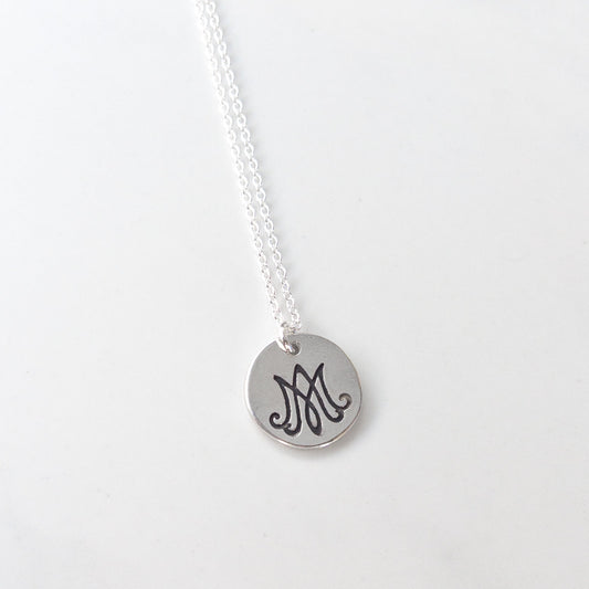 Close-up of round Marian monogram pendant in sterling silver, Auspice Maria symbol on white background. Necklace by Sagely Sparrow