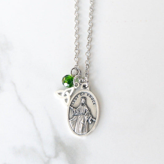 Silver-tone Catholic necklace featuring an Our Lady of Knock medal with the inscription “Pray for Us” on the back. The medal is paired with a silver triquetra (Trinity knot) charm and a small green glass bead, all hanging from a delicate silver chain.