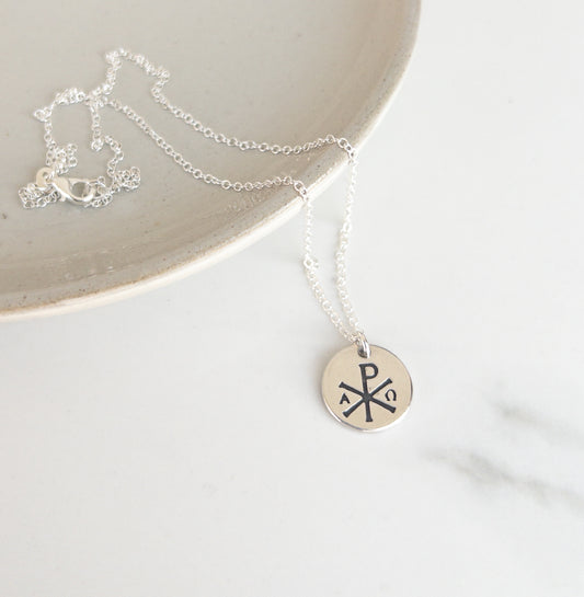 Silver necklace with a round pendant featuring thhe Chi Rho and Alpha and Omega symbols on a white surface