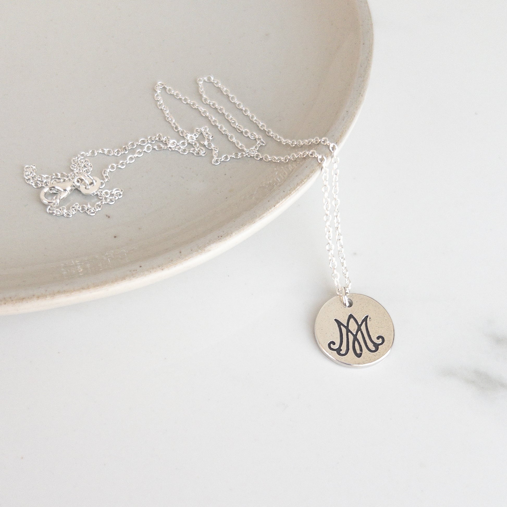 Silver necklace with a round pendant featuring letters "A" and "M" intertwined, for "Auspice Maria," on a white surface.