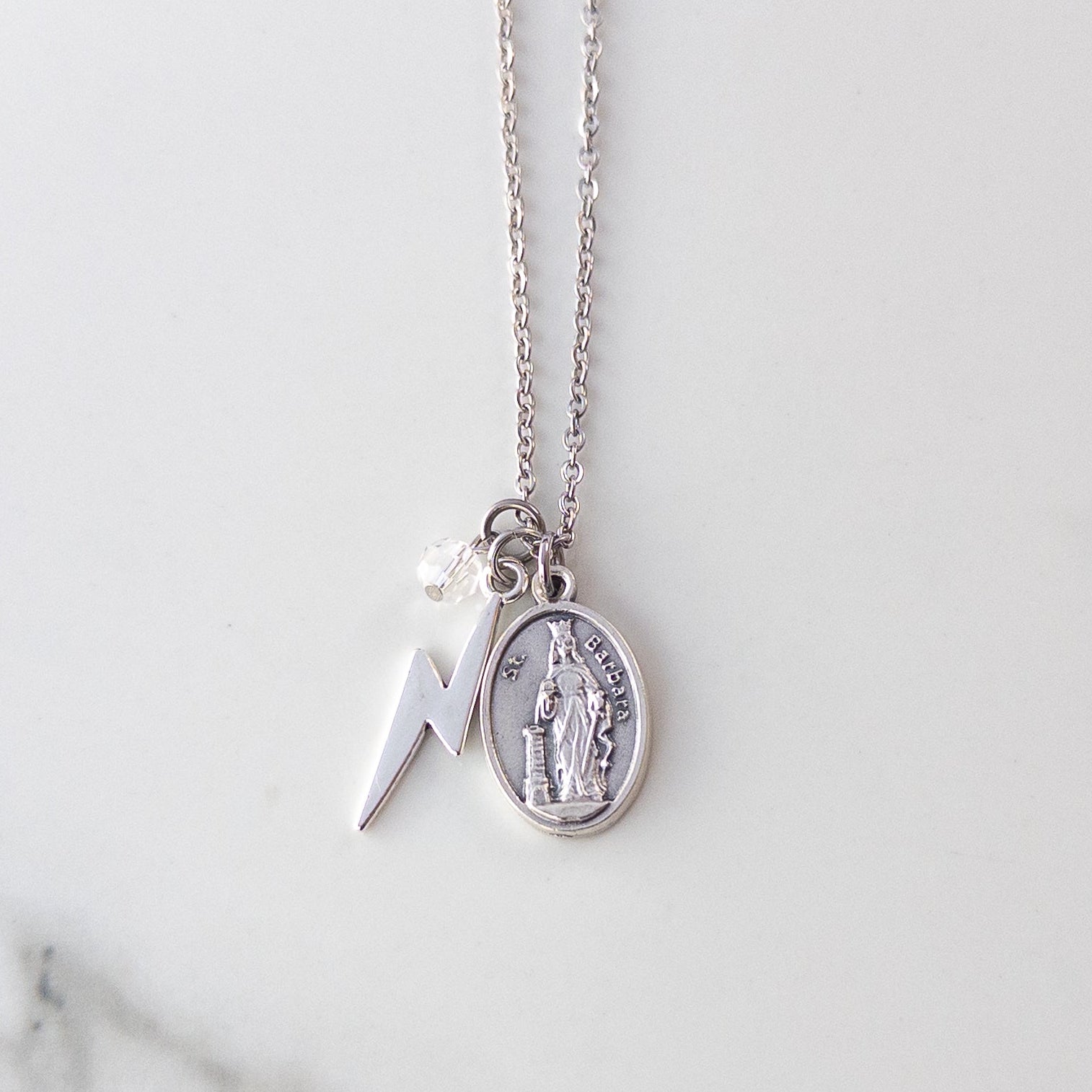 Silver-tone St Barbara necklace, featuring saint medal, lightning bolt, and clear bead on a white background