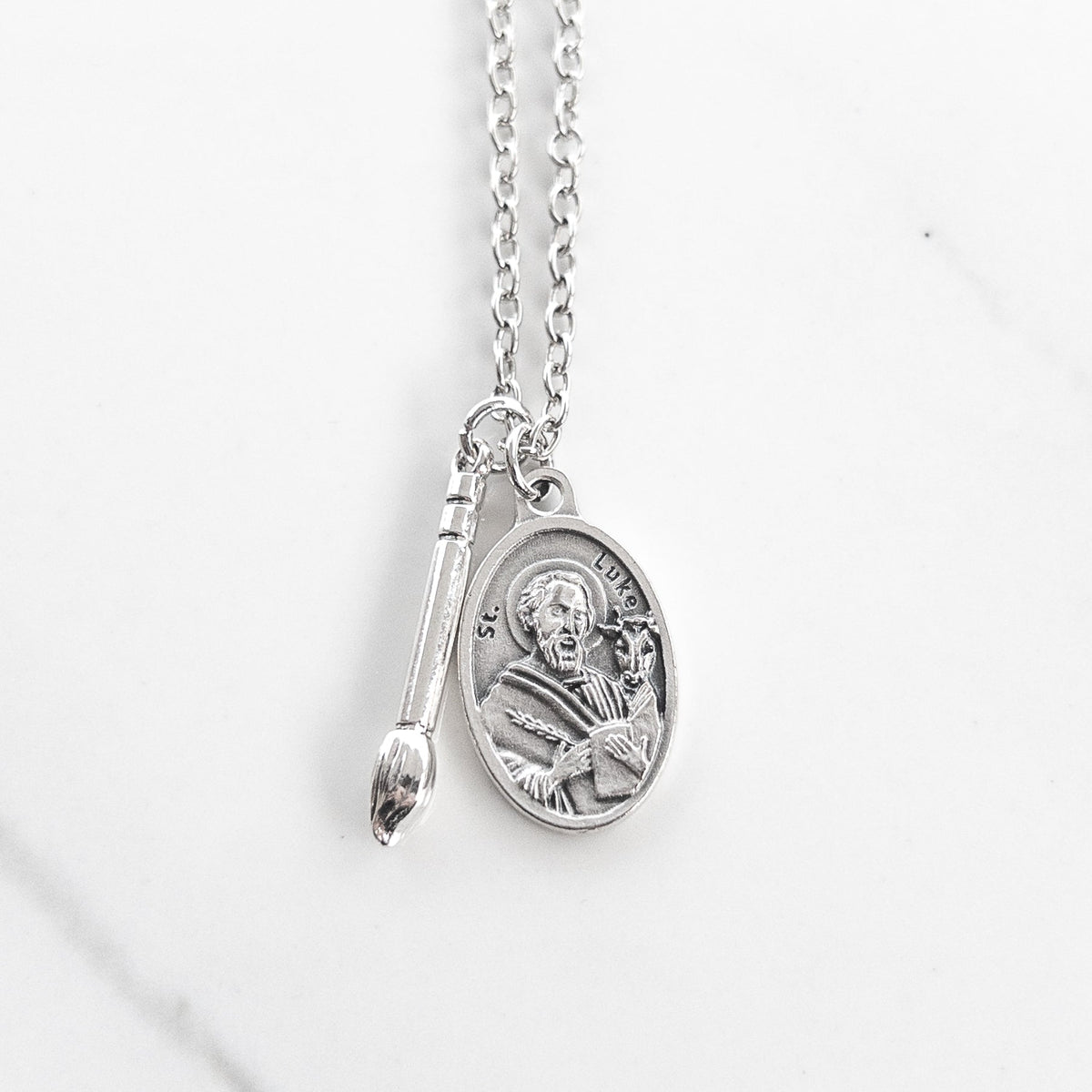 St. Luke necklace with a silvertone paintbrush charm and oval saint medal on a silver chain, photographed on white marble