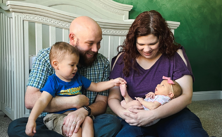 The Sagely Sparrow Family - a family of 4: father with toddler son on lap and mother with infant daughter in her arms, seated on floor in nursery