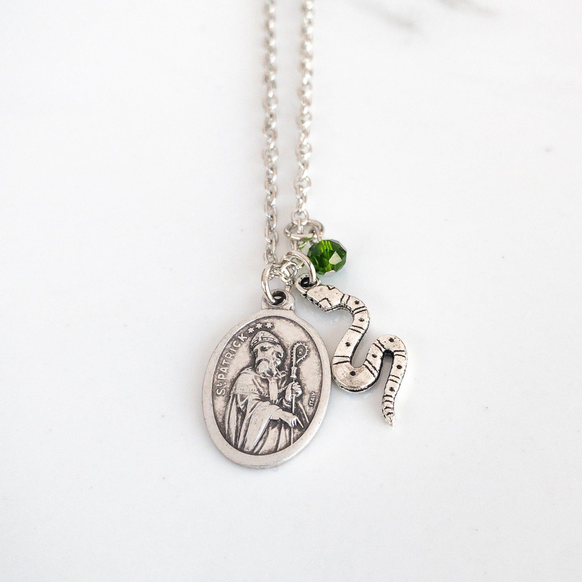 Silver tone necklace with St. Patrick medal, snake flat snake charm, and small green bead suspended from a steel chain on a white background
