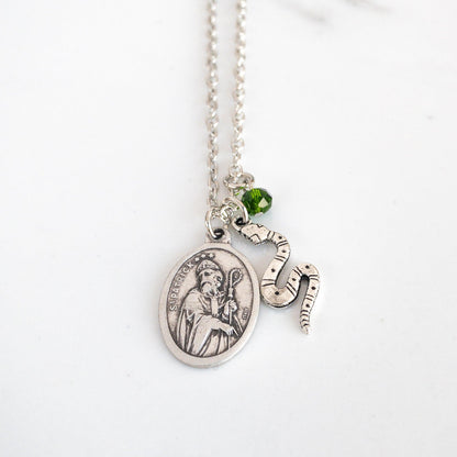 Silver tone necklace with St. Patrick medal, snake flat snake charm, and small green bead suspended from a steel chain on a white background