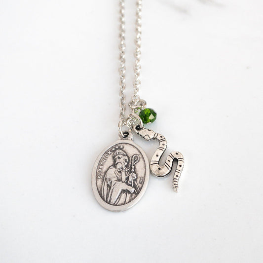 Silver tone necklace with St. Patrick medal, snake flat snake charm, and small green bead suspended from a steel chain on a white background